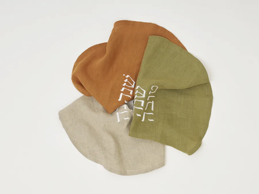 Three round challah covers in cinnamon, olive, and natural linen, embroidered with Shanah Tovah.